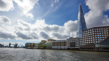 This is an urban landscape photograph captured on a summer morning, featuring London Bridge Hospital as the main subject. The view shows the hospital situated along the River Thames, with modern architecture prominent in the surrounding buildings. To the right, The Shard rises sharply into the sky, serving as a well-known London landmark and highlighting the contemporary architectural style of the area. The wide expanse of the Thames is visible in the foreground, and in the distance, the outline of Tower Bridge and HMS Belfast further emphasize the location's importance along the river. The overall scene showcases the blend of historic and modern architecture typical of central London, with soft morning light illuminating the buildings and clouds overhead.